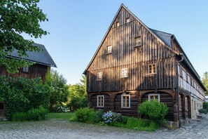 Suuri puinen rakennus, jossa on useita ikkunoita ja kalteva katto. Sen ympärillä on vehreää kasvillisuutta ja mukulakivipolku.