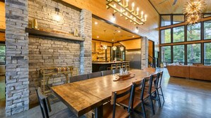 A spacious dining area with a long wooden table and chairs, a stone accent wall, and a kitchen in the background.