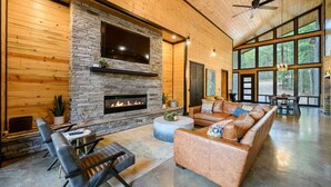 A spacious living room with a stone fireplace, leather sofa, and large windows offering a view of the outdoors.