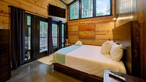A wooden cabin bedroom with a bed, a painting, and a large window overlooking a forest.