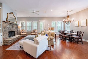 A spacious living room with a stone fireplace, a white sofa, a dining area with a wooden table and chairs, and large windows with blinds.