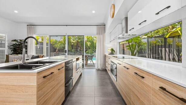 A modern kitchen with a central island, wooden cabinets, and a view of a pool and greenery outside. - The Palms on Nautilus (Kingscliff)