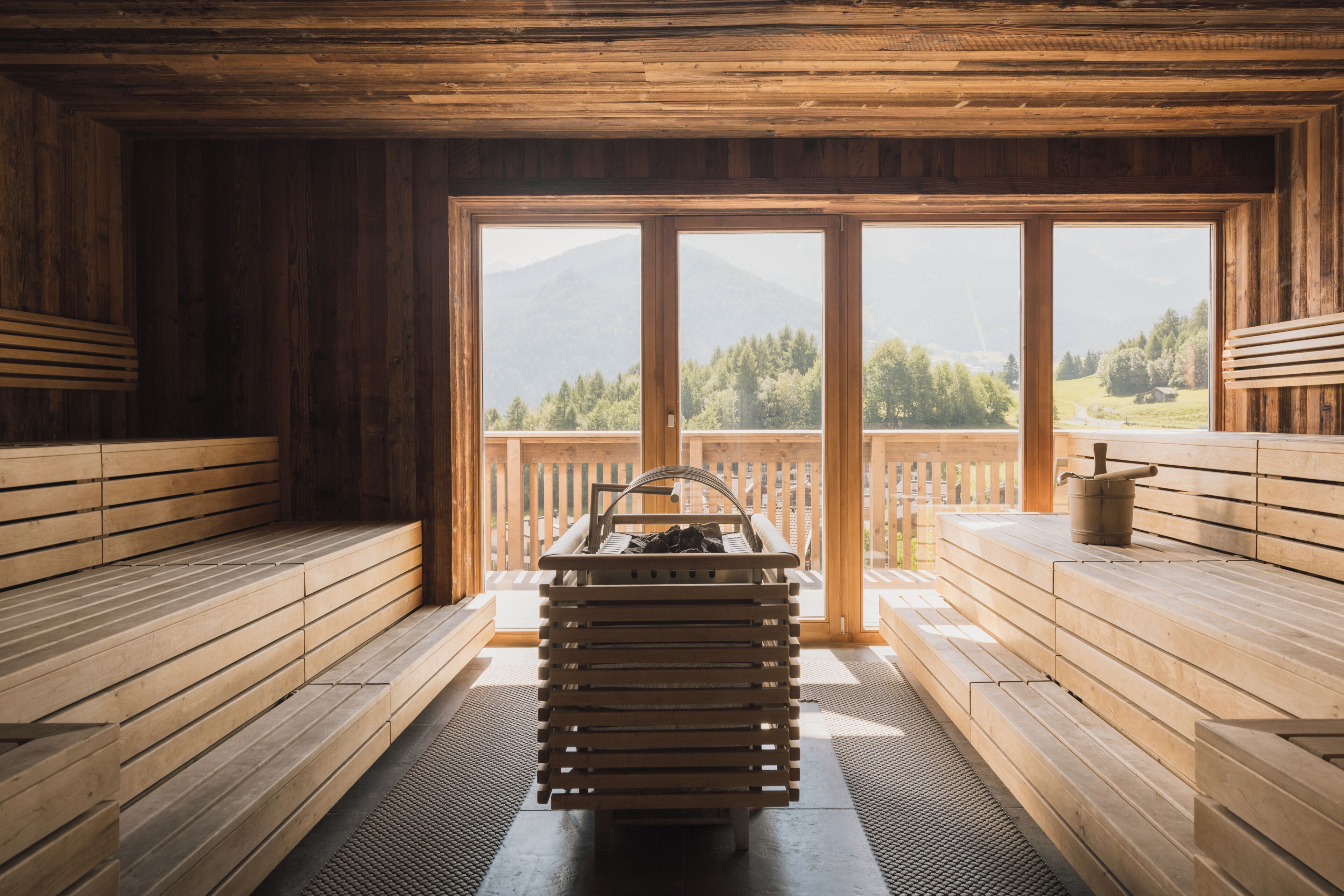 Eine Holzsauna mit Bänken, eine Bank mit Eimer und Blick auf Berge und Bäume durch große Fenster.