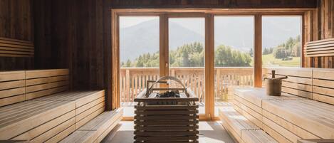 Eine Holzsauna mit Bänken, eine Bank mit Eimer und Blick auf Berge und Bäume durch große Fenster.