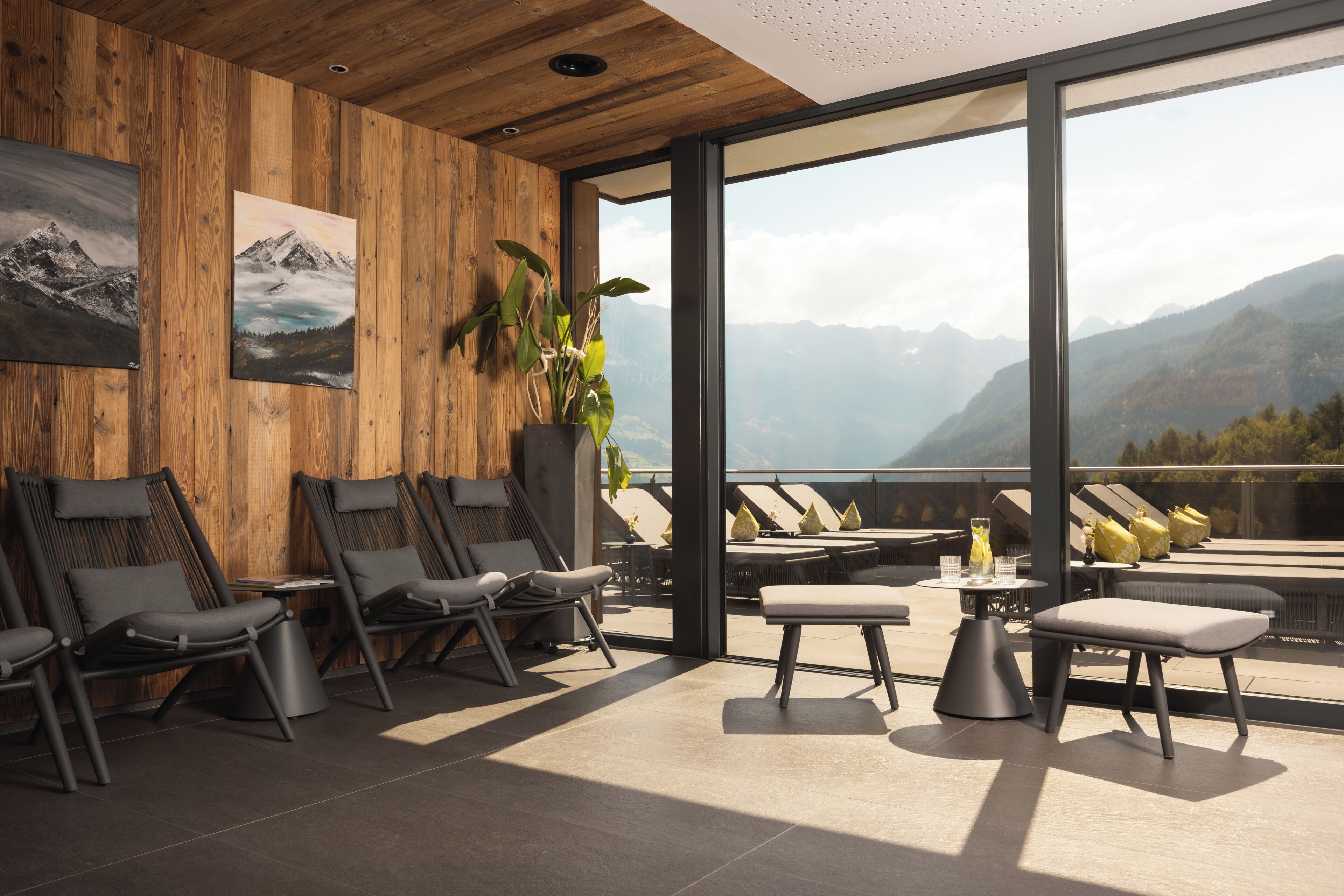 A modern lounge area with wooden paneling, reclining chairs, a potted plant, and a view of mountains through large windows.
