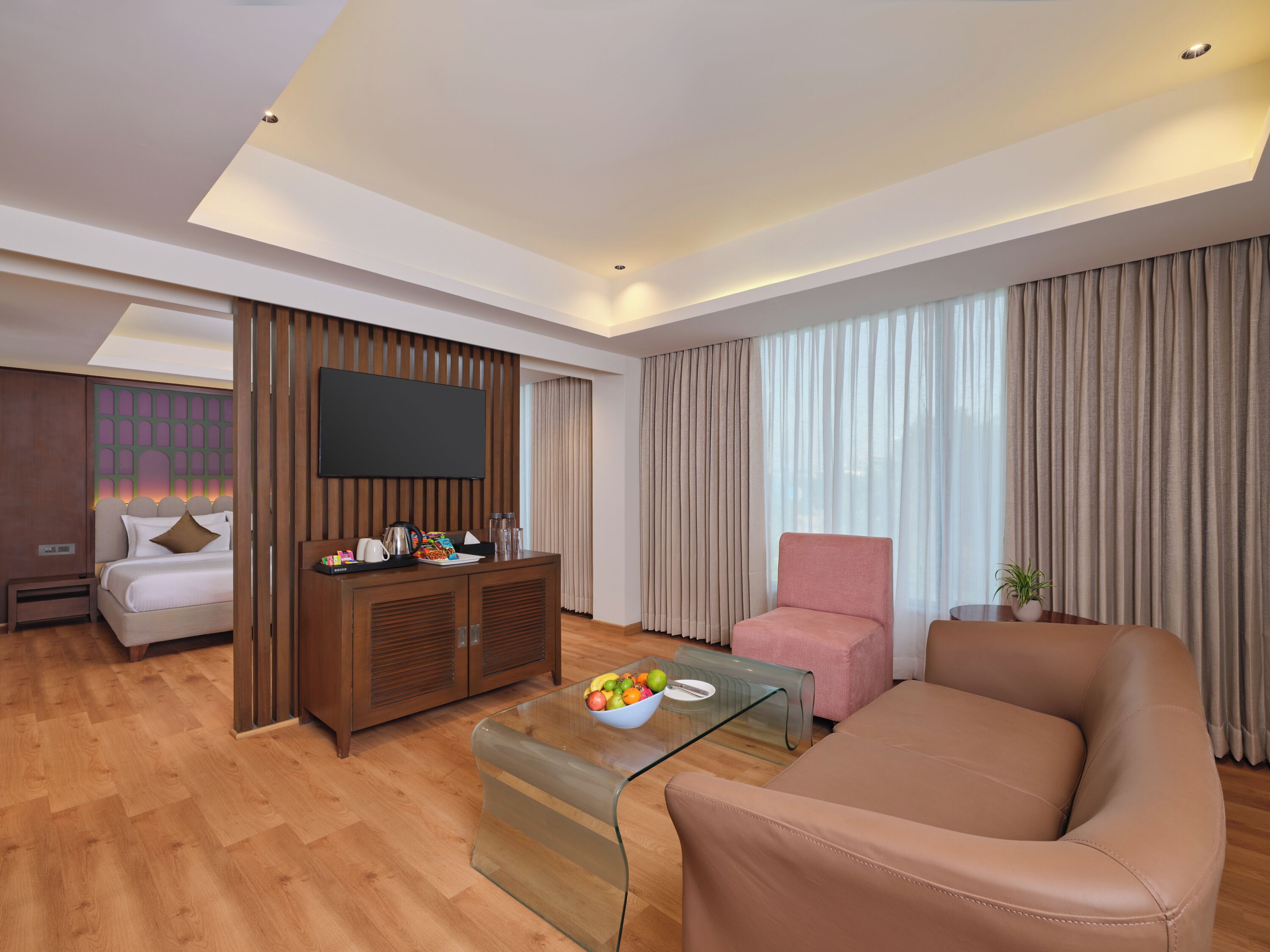 Una camera d'albergo moderna con un letto, una TV, un tavolino da caffè in vetro con frutta, una poltrona rosa e un divano.
