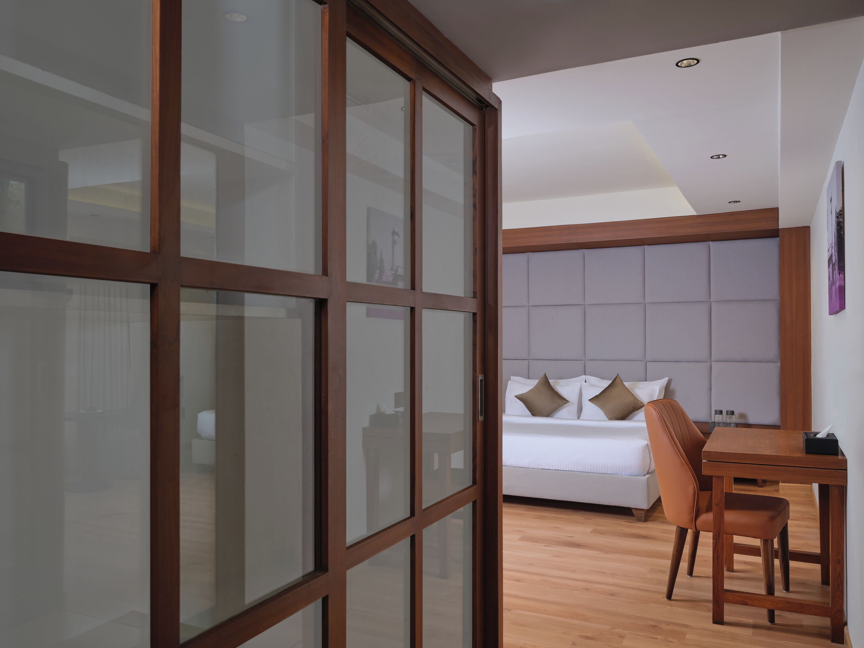 A modern hotel room with a wooden floor, a bed with white bedding, a wooden dining table, and a large sliding glass door.