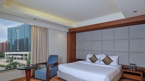 A hotel room with a large bed, a chair, a small table with a plate of food, and a view of a cityscape. - Regenta Place Ahmedabad (Ahmedabad)