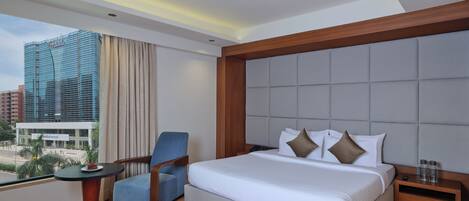 A hotel room with a large bed, a chair, a small table with a plate of food, and a view of a cityscape.