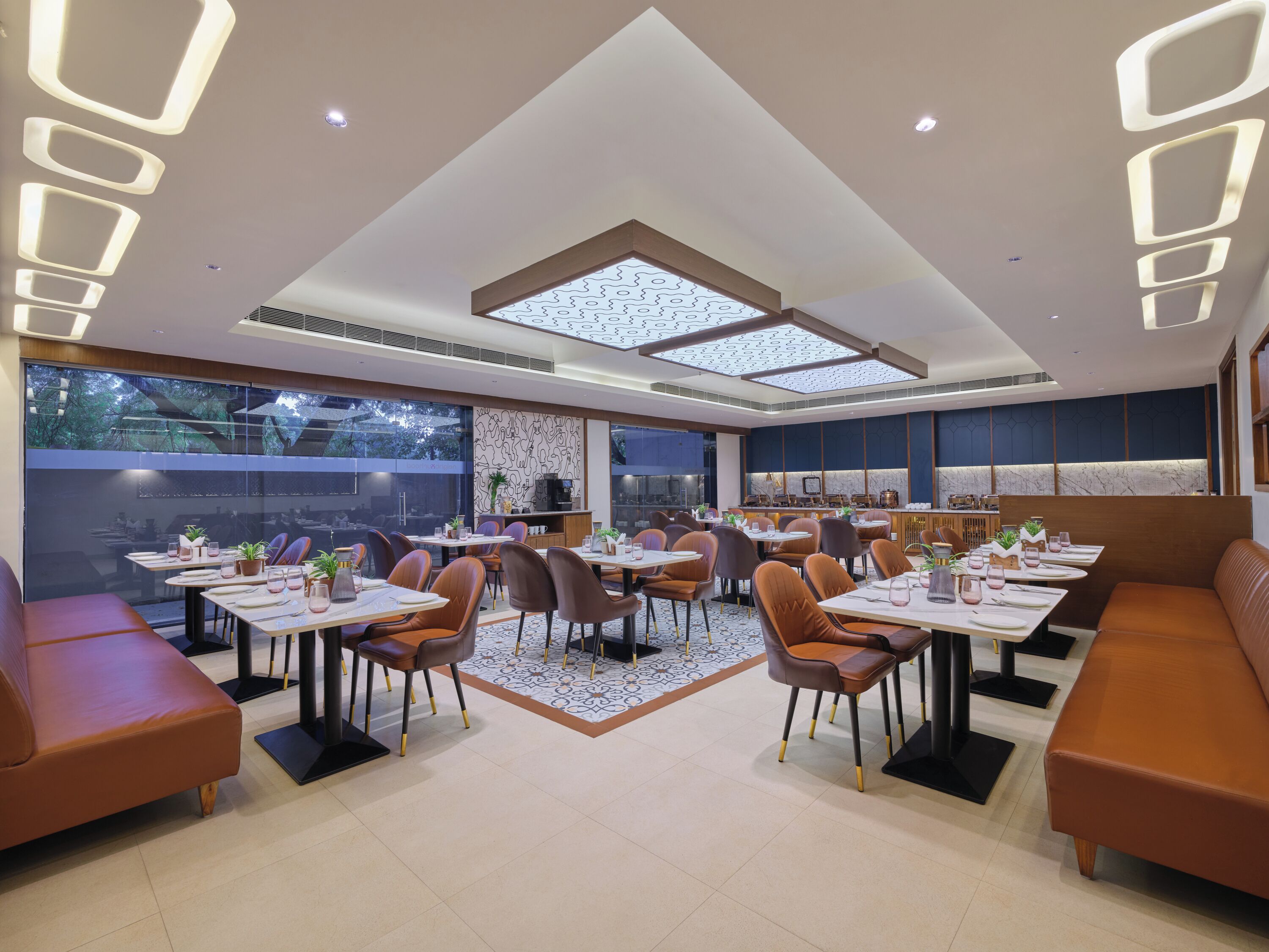 A modern dining area with high ceilings, recessed lighting, and a central skylight. Tables are set with chairs and centerpieces.