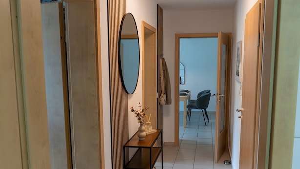 A hallway with a wooden floor, a mirror, a small table with a vase, and a chair in the background.
