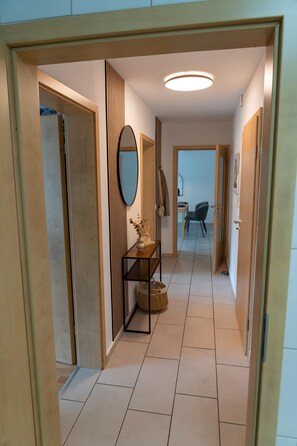 A hallway with a wooden floor, a mirror, a small table with a vase, and a chair in the background. - Apartment 'Retreat' with Mountain View, Private Garden and Wi-Fi (Plettenberg)
