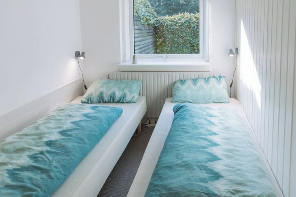 Une petite pièce lumineuse, avec deux lits simples, une fenêtre donnant sur un espace verdoyant et un radiateur en dessous.