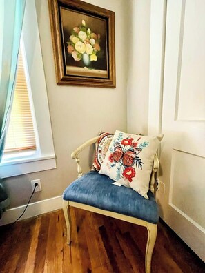 A corner room with a wooden floor, a white wall, a framed floral painting, and a blue cushioned chair with floral pillows.