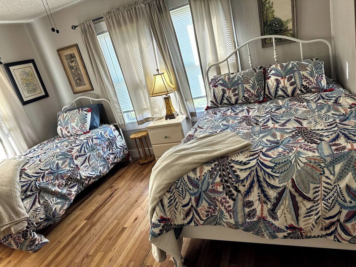 A bedroom with a double bed, a single bed, a bedside table, a lamp, and curtains.