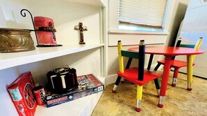 Une pièce avec une étagère blanche sur laquelle se trouvent une bougie rouge, une petite croix en bois, un jeu de société Monopoly et une boîte rouge. On y voit également une chaise rouge et jaune, avec une table rouge placée à côté.