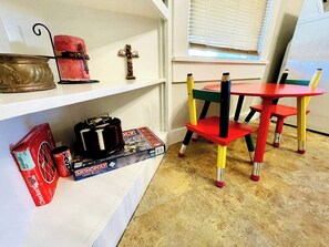 Ein Zimmer mit einer weißen Ablage, auf der eine rote Kerze, ein kleines hölzernes Kreuz, ein Monopoly-Spiel und eine rote Schachtel stehen. Daneben steht ein roter und gelber Stuhl und ein roter Tisch.