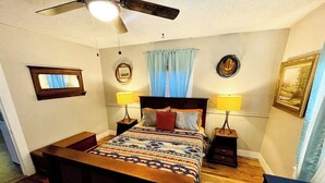 Une chambre à coucher avec un lit, deux tables de chevet, un ventilateur de plafond et une fenêtre avec des rideaux.
