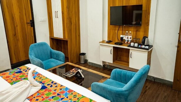 Une chambre d’hôtel comprenant un lit, deux fauteuils bleus, une petite table basse, un téléviseur fixé sur un panneau en bois, un mini-bar et une poubelle.