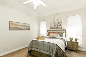 Un dormitorio con cama, mesitas de noche, ventilador de techo, obras de arte y una ventana con persianas.