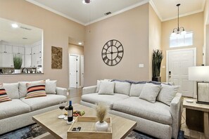 A modern living room with two sofas, a coffee table, a wall clock, and a kitchen in the background.