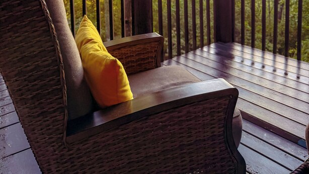 Une terrasse en bois, meublée de mobilier en osier et agrémentée d’un coussin jaune, offre une vue sur un paysage boisé avec des montagnes à l’horizon.
