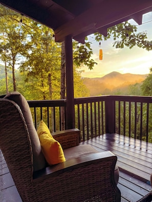 Une terrasse en bois, meublée de mobilier en osier et agrémentée d’un coussin jaune, offre une vue sur un paysage boisé avec des montagnes à l’horizon.
