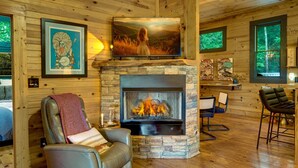 L’intérieur chaleureux d’un chalet avec une cheminée en pierre, un téléviseur fixé au mur et des œuvres d’art encadrées qui ornent les murs.