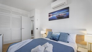 Ein ordentlich bezogenes Bett mit einer blauen Steude, ein Bild eines Bootes an der Wand und eine weiße Klimaanlage darüber.