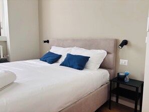 Una cama bien hecha con almohadas blancas y azules, un cabecero color beige y una mesita de noche con una lámpara y una botella de agua.