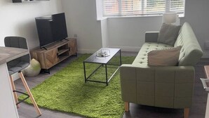 A living room with a green sofa, a coffee table, a TV on a wooden stand, and a window with blinds.