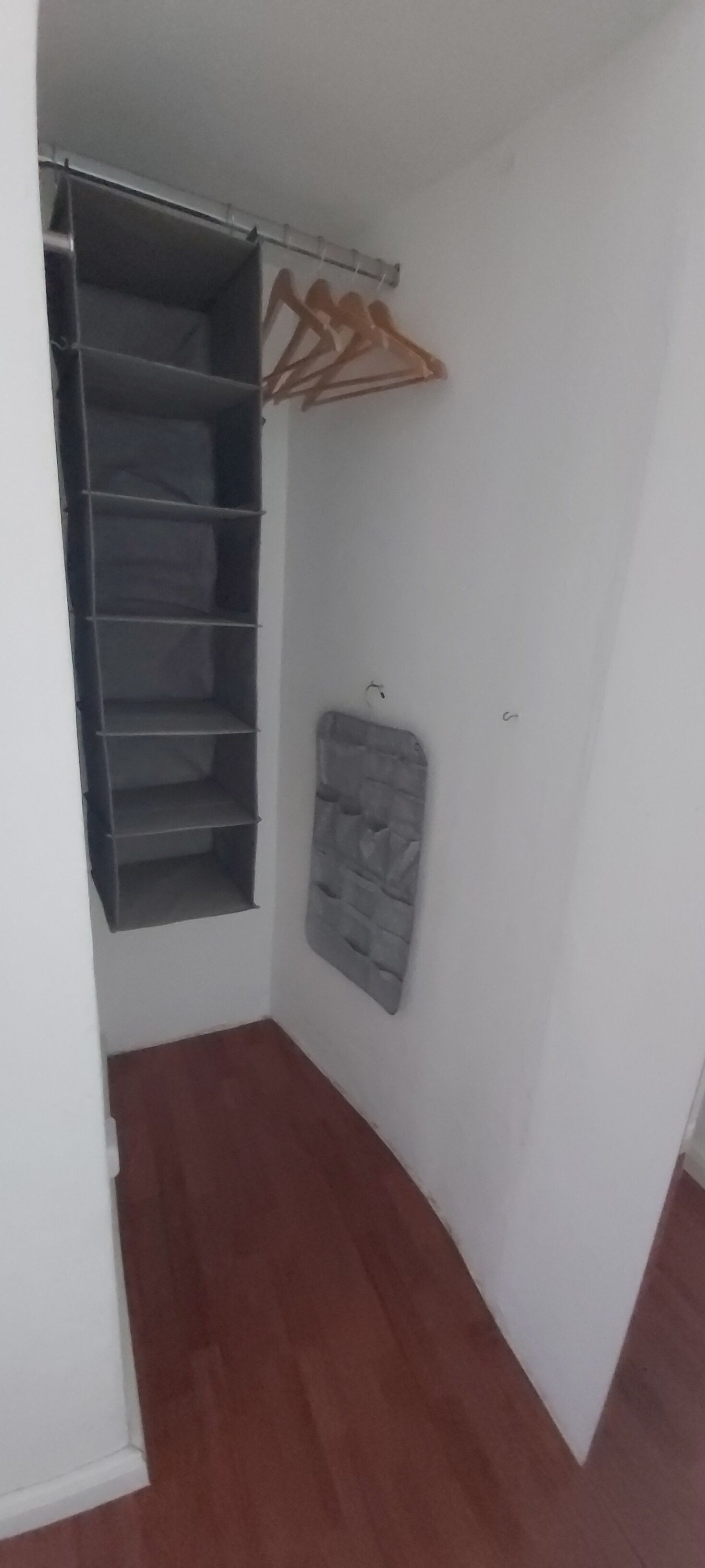 Ein Eckbereich eines Zimmers mit integriertem Kleiderschrank, einer grau gepolsterten Bank und einem Holzkleiderständer.