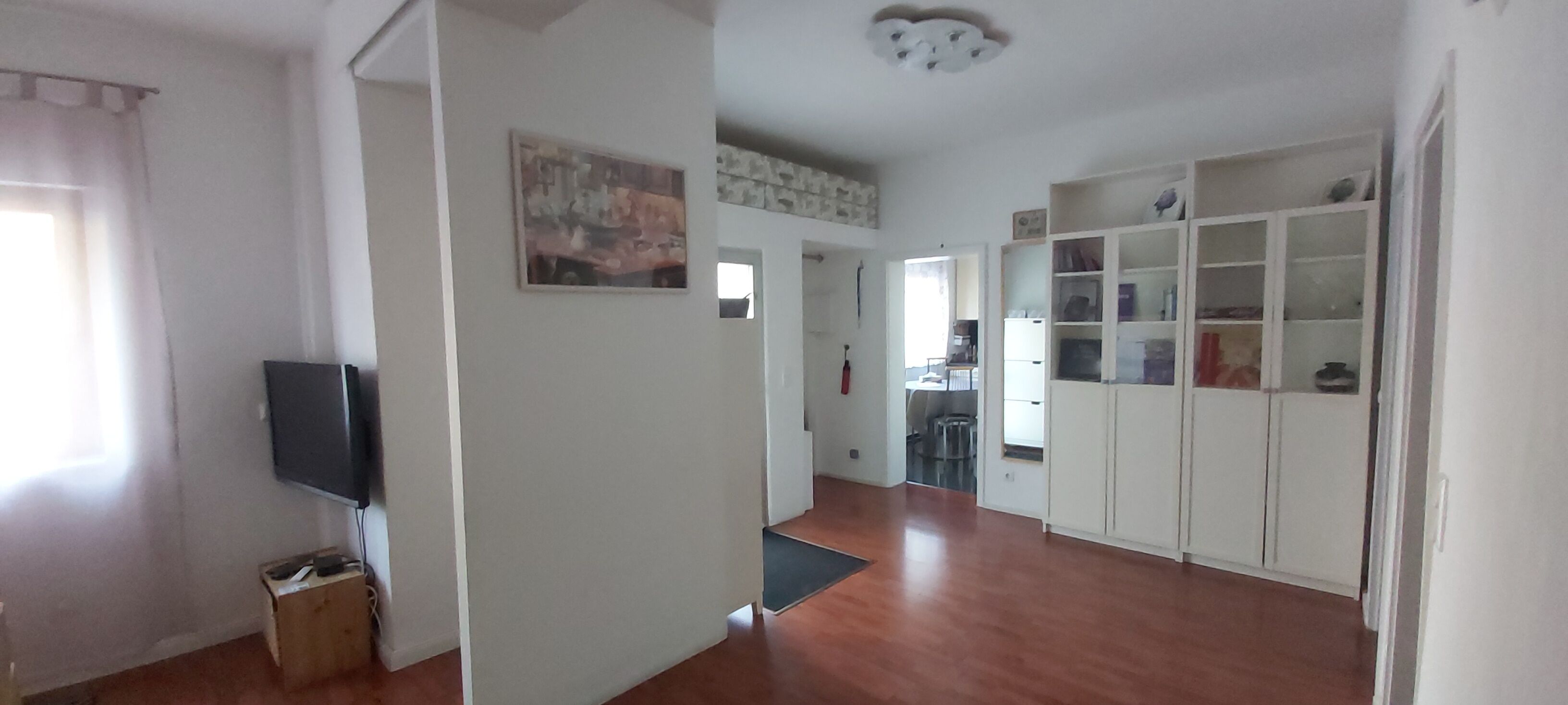 A spacious living area with a wooden floor, a flat-screen TV mounted on the wall, and a bookshelf filled with various items.