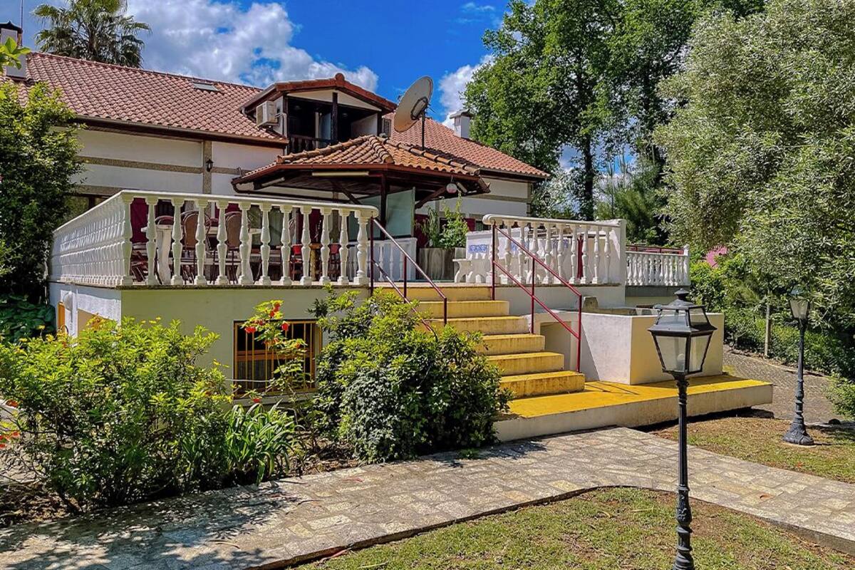 A two-story house with a red-tiled roof, a balcony with white railings, and a small garden with a pathway leading to the entrance.