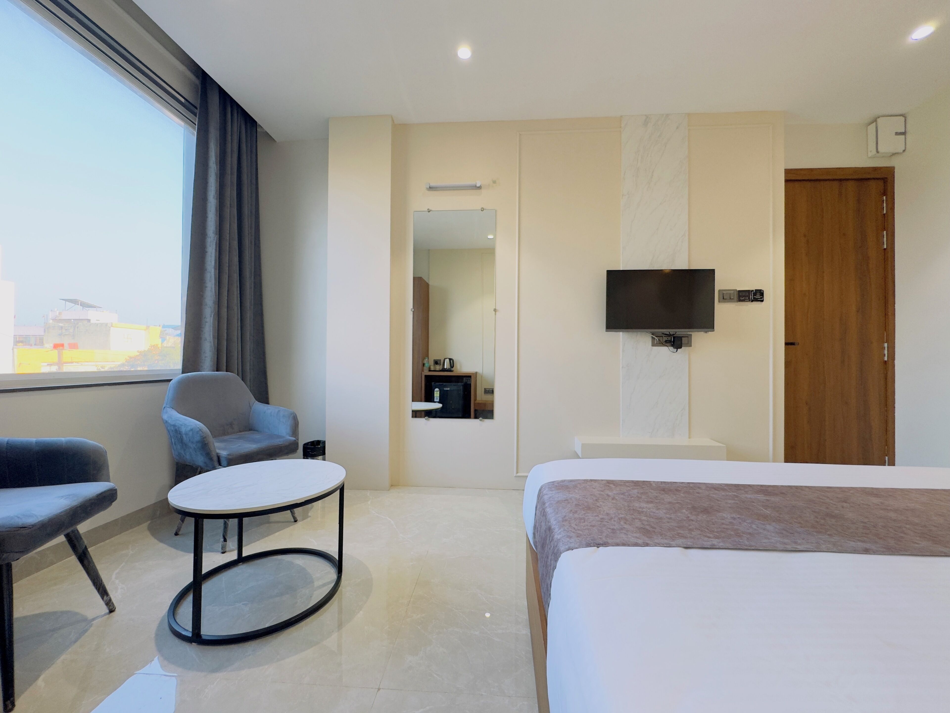 A hotel room with a bed, a TV mounted on the wall, a small table, two chairs, a mirror, and a window with curtains.