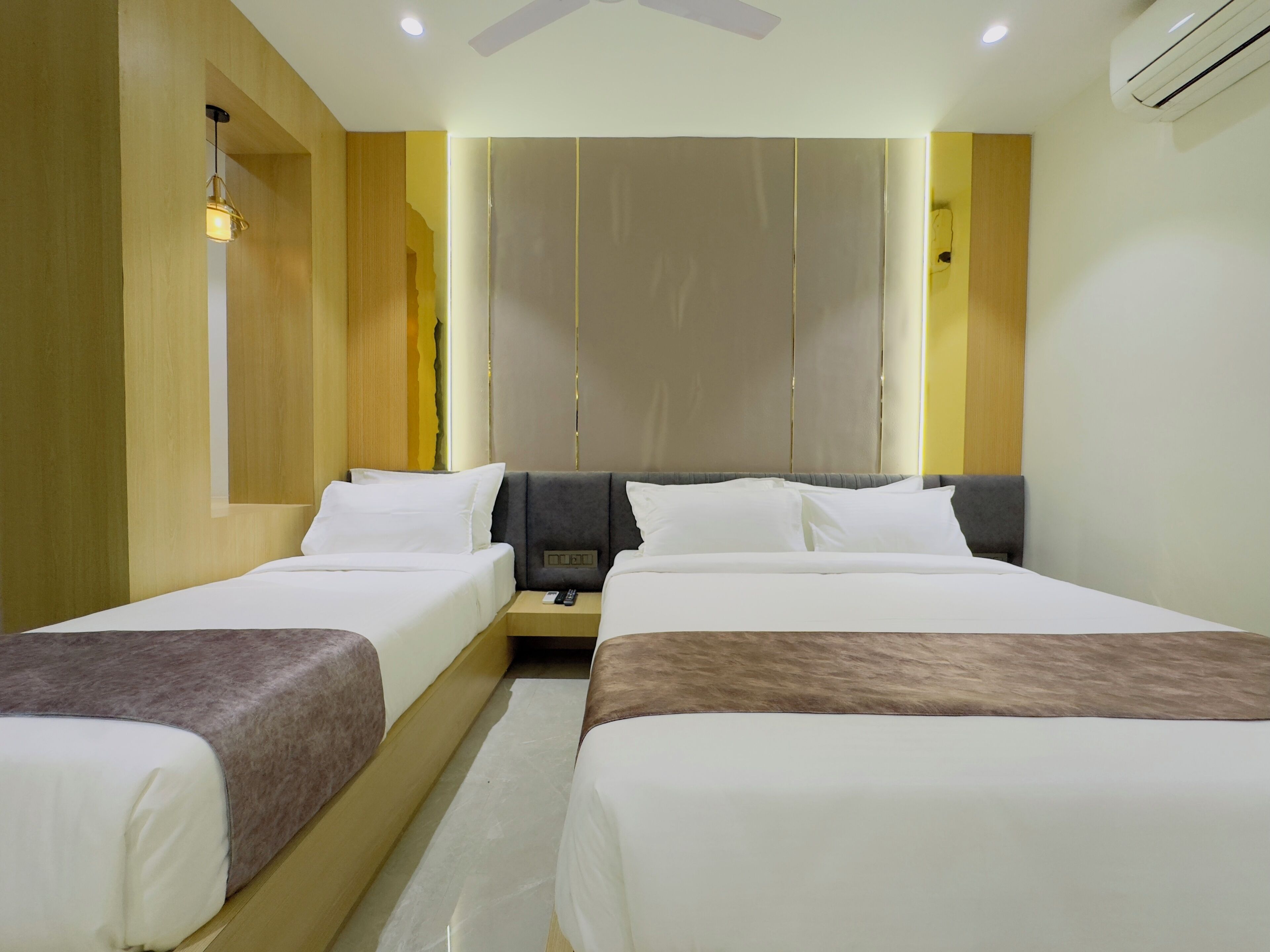 A hotel room with two beds, a ceiling fan, an air conditioning unit, and a bedside table with a remote control.