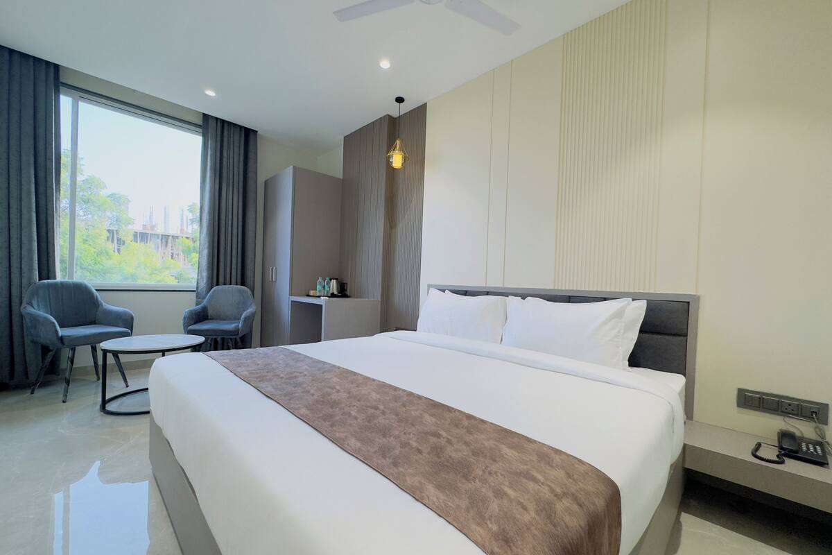 Une chambre d’hôtel avec un grand lit, deux fauteuils, une petite table, un bureau avec un téléphone et une fenêtre avec des rideaux.