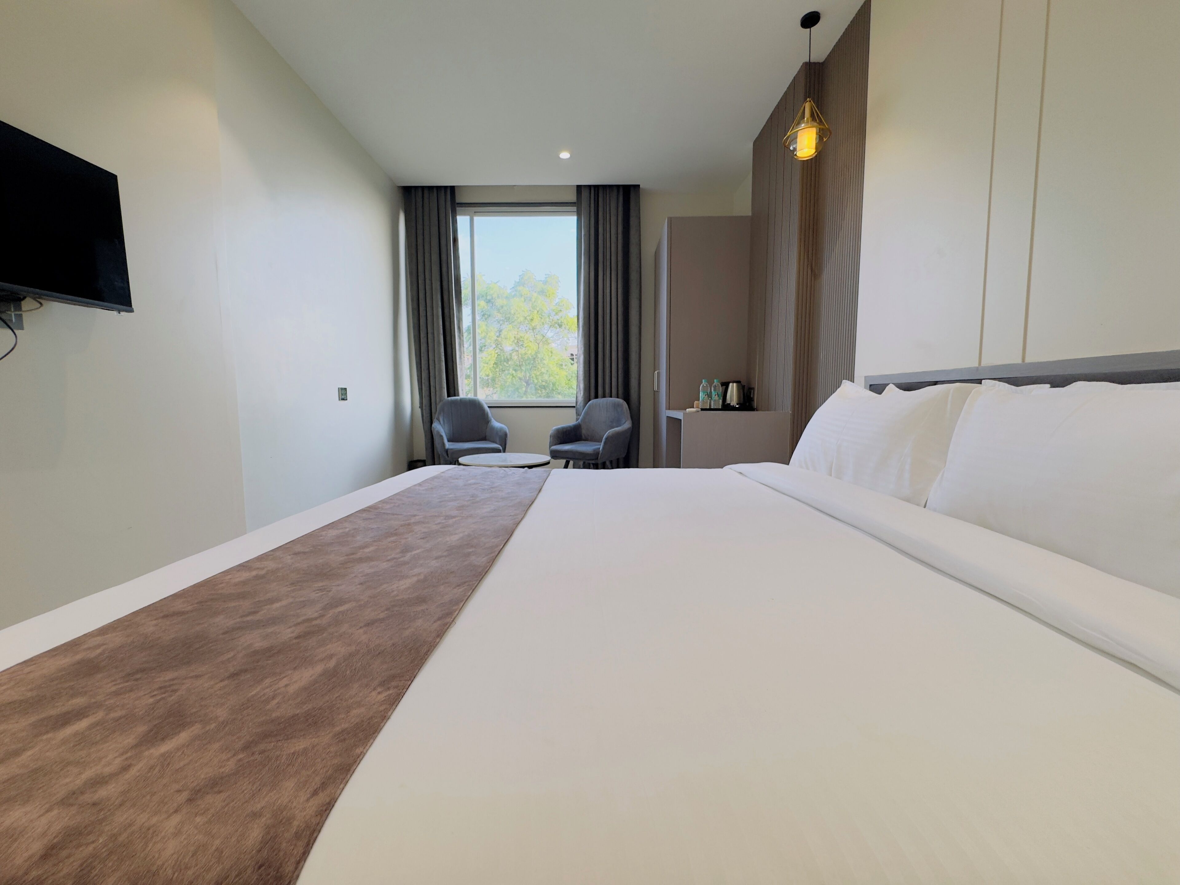 A hotel room with a large bed, two armchairs, a small table, a TV mounted on the wall, and a window with curtains.