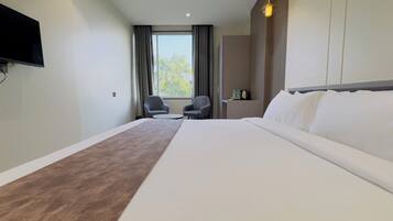 A hotel room with a large bed, two armchairs, a small table, a TV mounted on the wall, and a window with curtains.