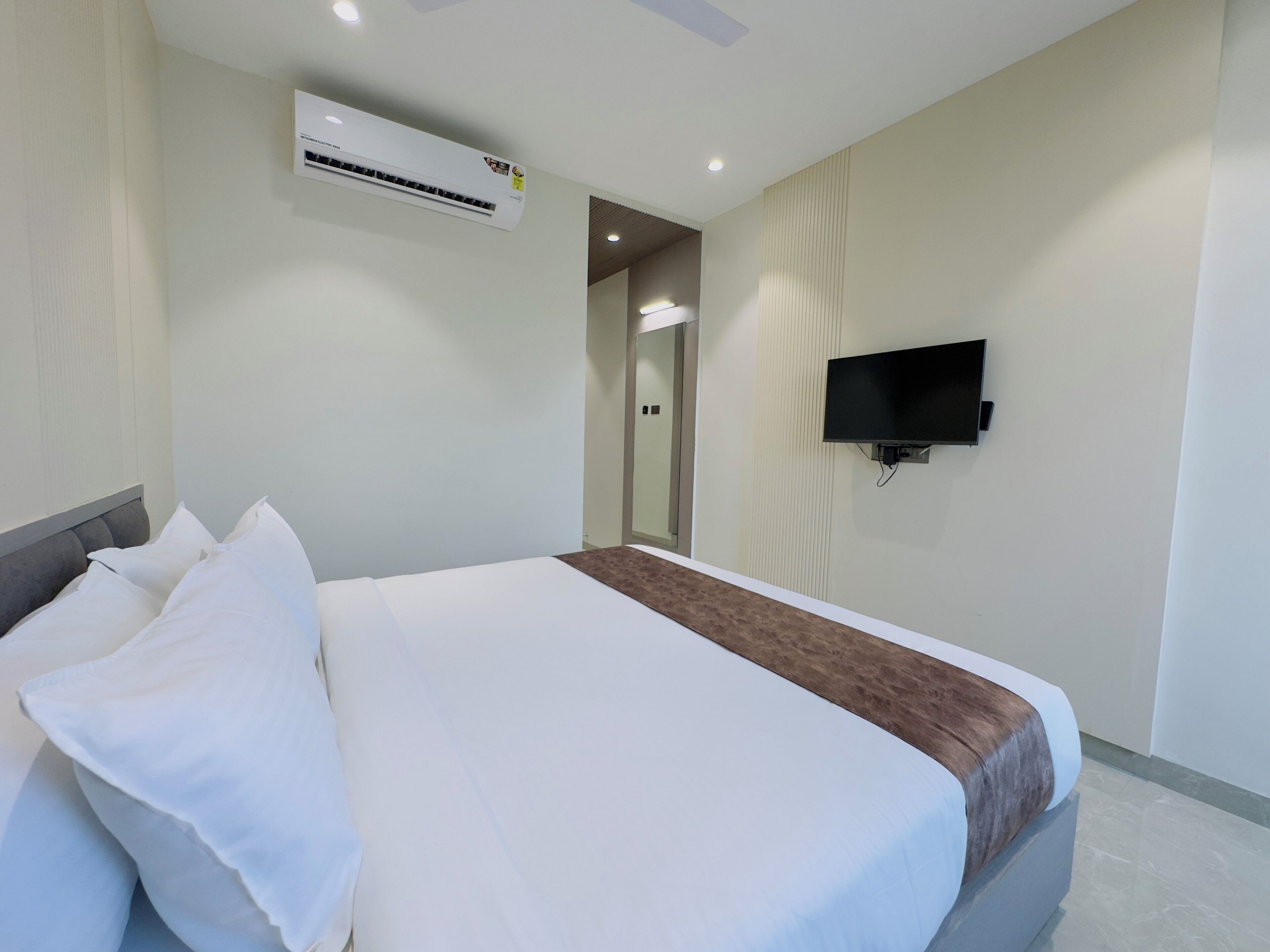 A hotel room with a bed, a flat-screen TV mounted on the wall, and an air conditioning unit above the bed.