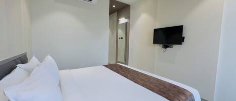 A hotel room with a bed, a flat-screen TV mounted on the wall, and an air conditioning unit above the bed.