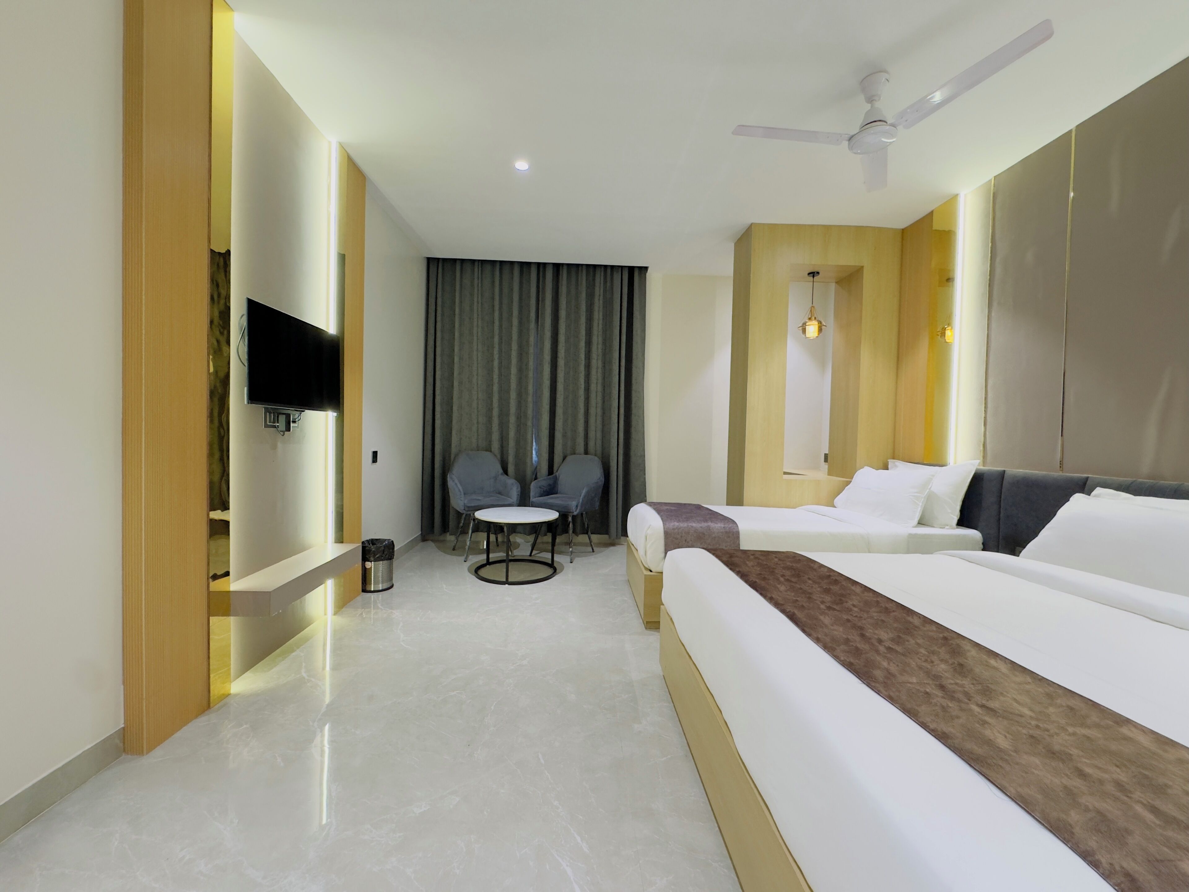 A hotel room with a bed, a TV, a ceiling fan, a small table with two chairs, and a curtain.