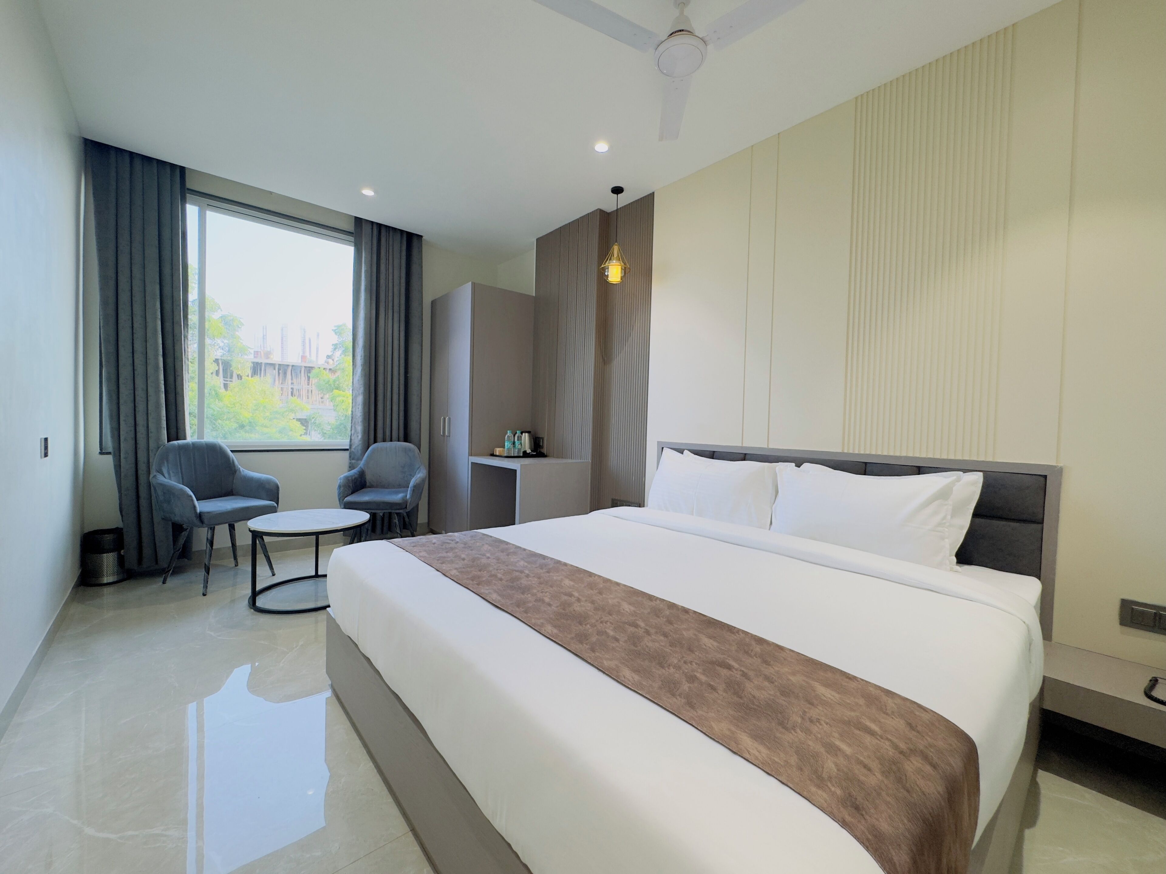 A modern hotel room with a large bed, a desk, two chairs, and a window with curtains.