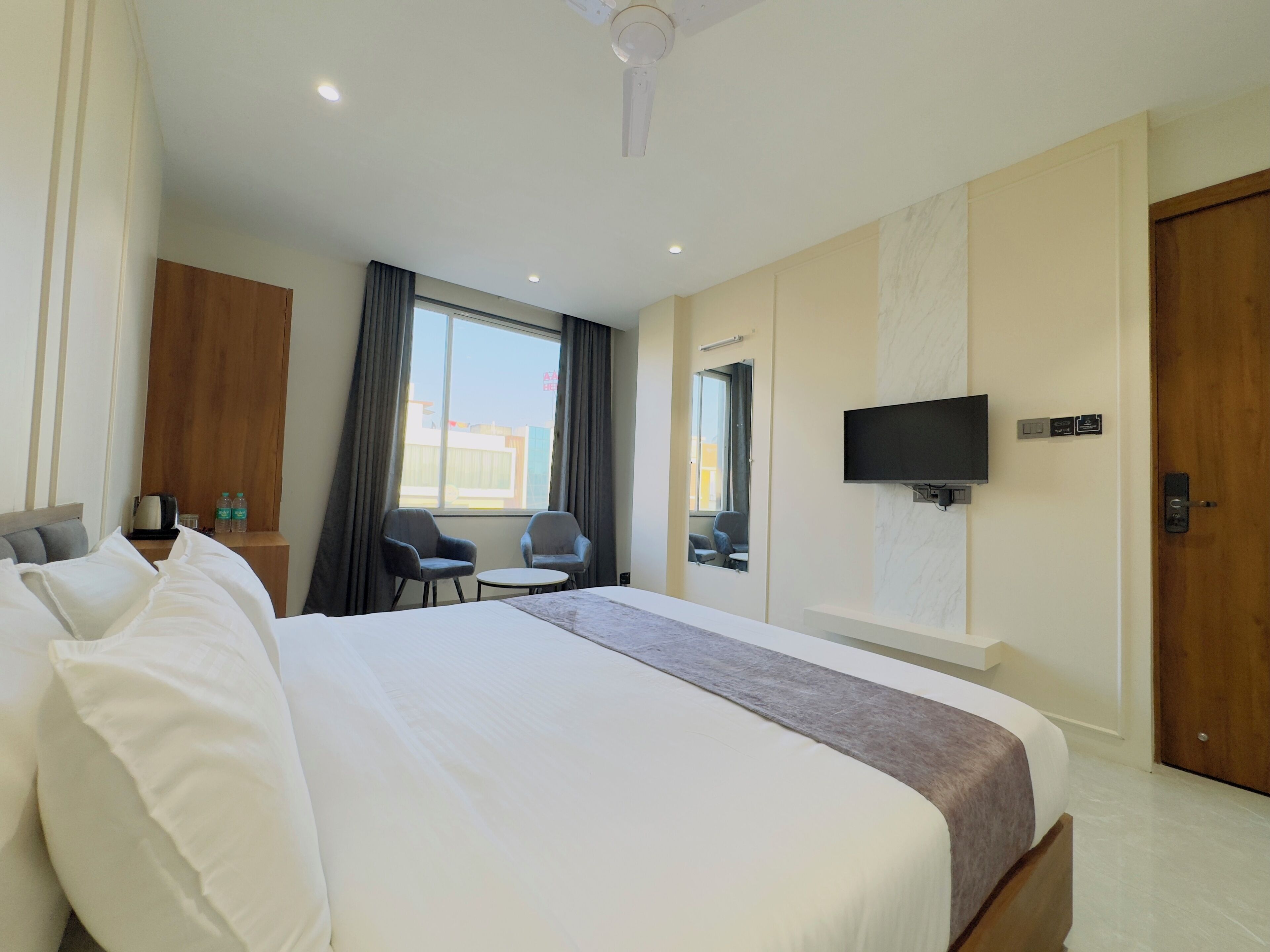 A hotel room with a large bed, two armchairs, a small table, a TV mounted on the wall, and a window with curtains.