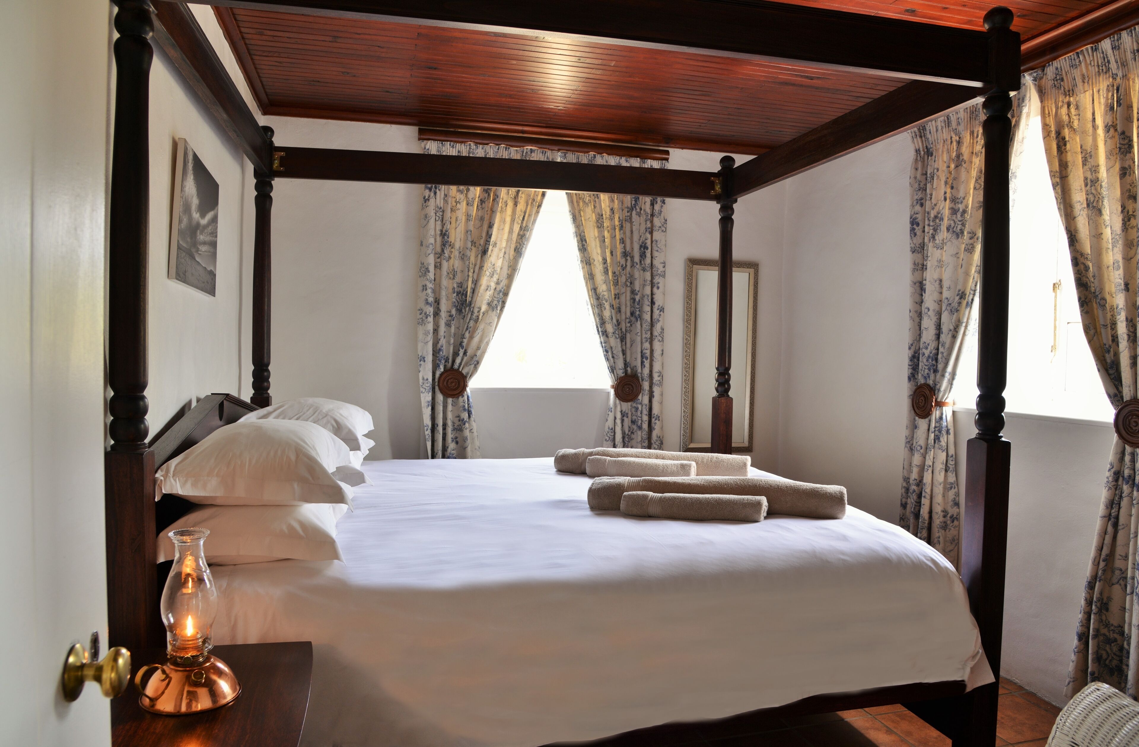 A four-poster bed with white linens and rolled towels, a brass door handle, and a lit candle on a wooden nightstand.