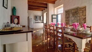A dining area with wooden chairs and a table set for a meal, a kitchen in the background, and a stone wall. - Melkkamer by De Hoop Collection (Overberg)