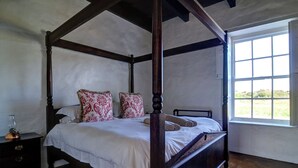 A four-poster bed with white linens and patterned pillows, a wooden nightstand with a glass bottle, and a window with a view of greenery. - Melkkamer by De Hoop Collection (Overberg)