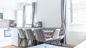 Une cuisine moderne avec des meubles blancs, un coin repas doté d’un banc capitonné et de quatre chaises, ainsi qu’une grande fenêtre agrémentée de rideaux.