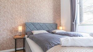 Une chambre avec un papier peint à motifs, un lit avec une literie grise, une table de chevet avec une lampe et une fenêtre avec des rideaux.