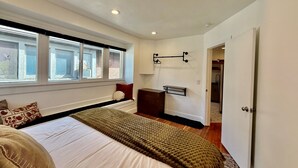 Une chambre avec un grand lit, une fenêtre avec des stores, un luminaire fixé au mur et une porte donnant sur une salle de bain.